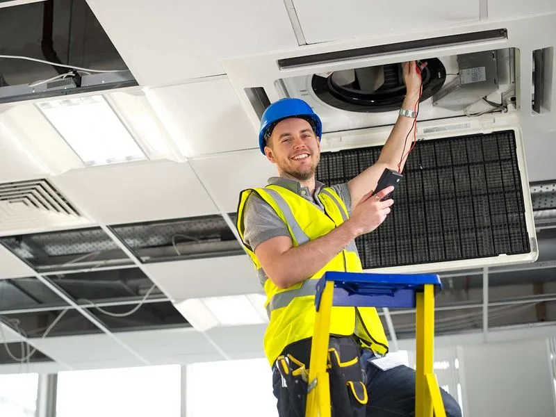 Techniker mit Schutzhelm und Warnweste überprüft eine Klimaanlage an der Decke.